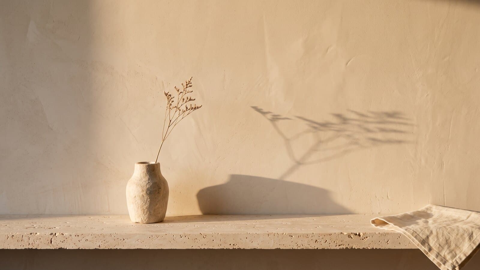 Morning light falling across a warm plaster wall and a travertine ledge, a dried stem in a small stone vase casting a long shadow.