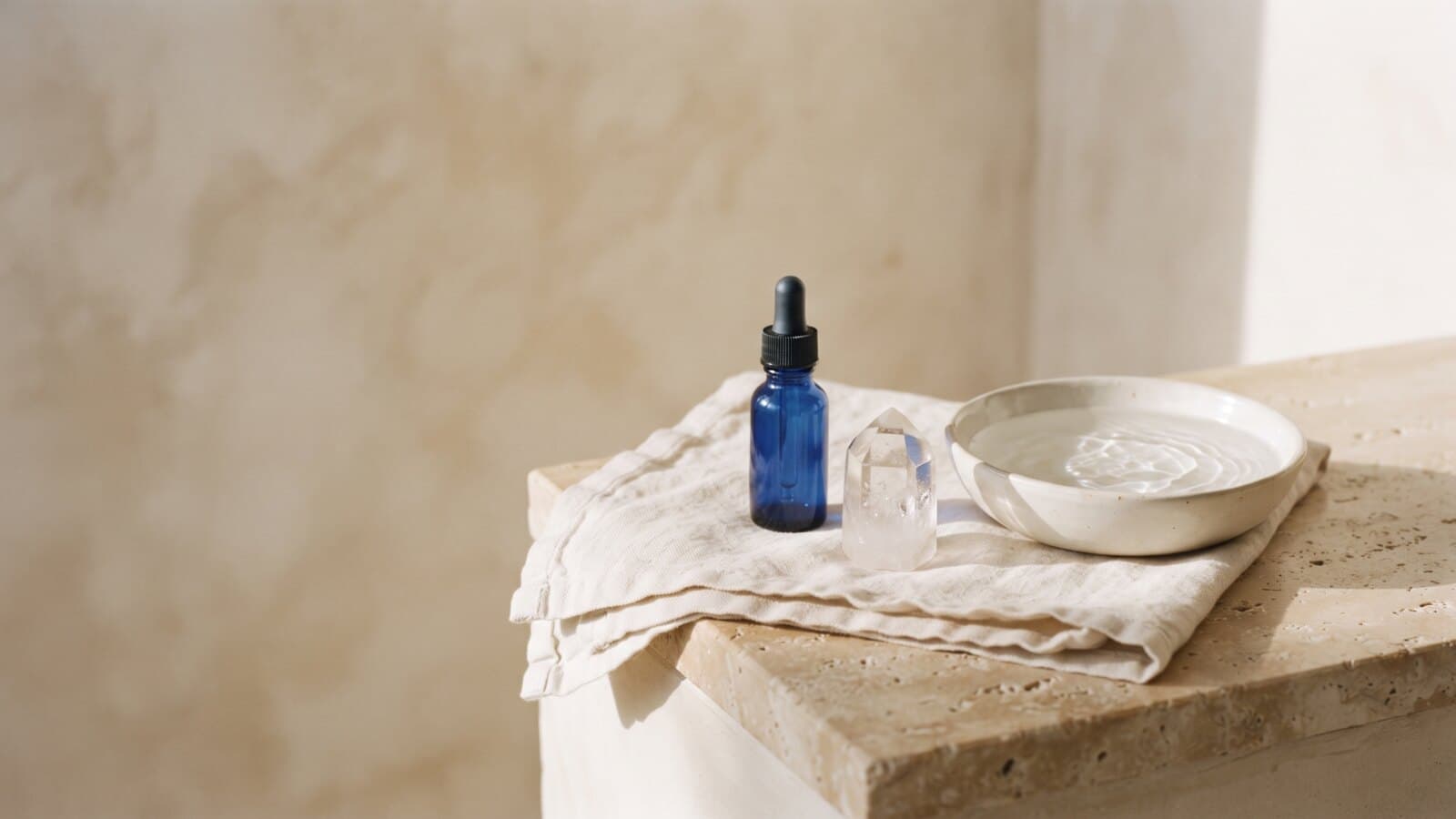 A shallow dish of clear water, a faceted quartz crystal and a small unlabeled cobalt-blue dropper bottle on a stone ledge in warm side light.