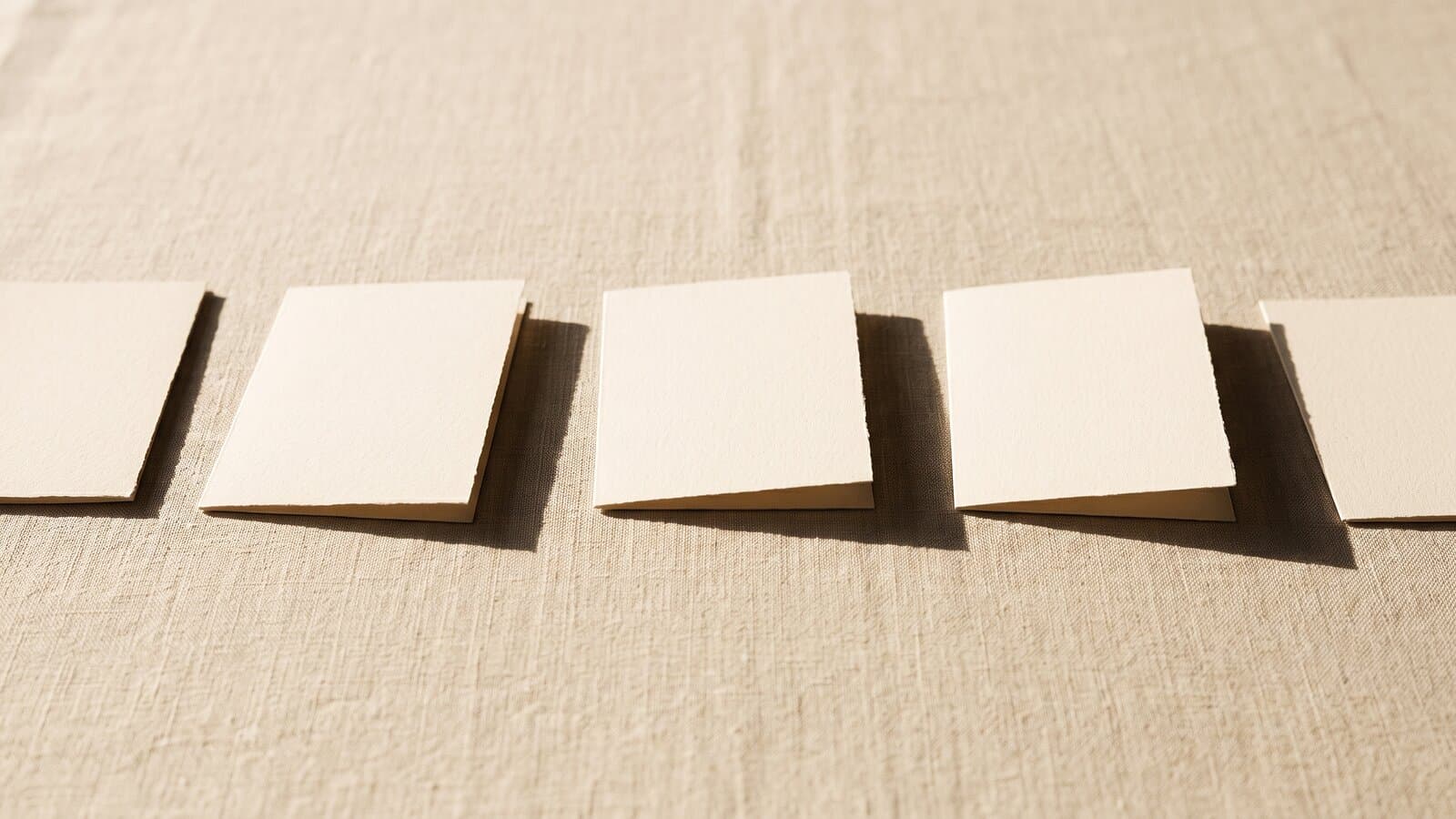 A row of small blank cream cards laid left to right across pale linen in low raking sunlight, each throwing a long shadow.