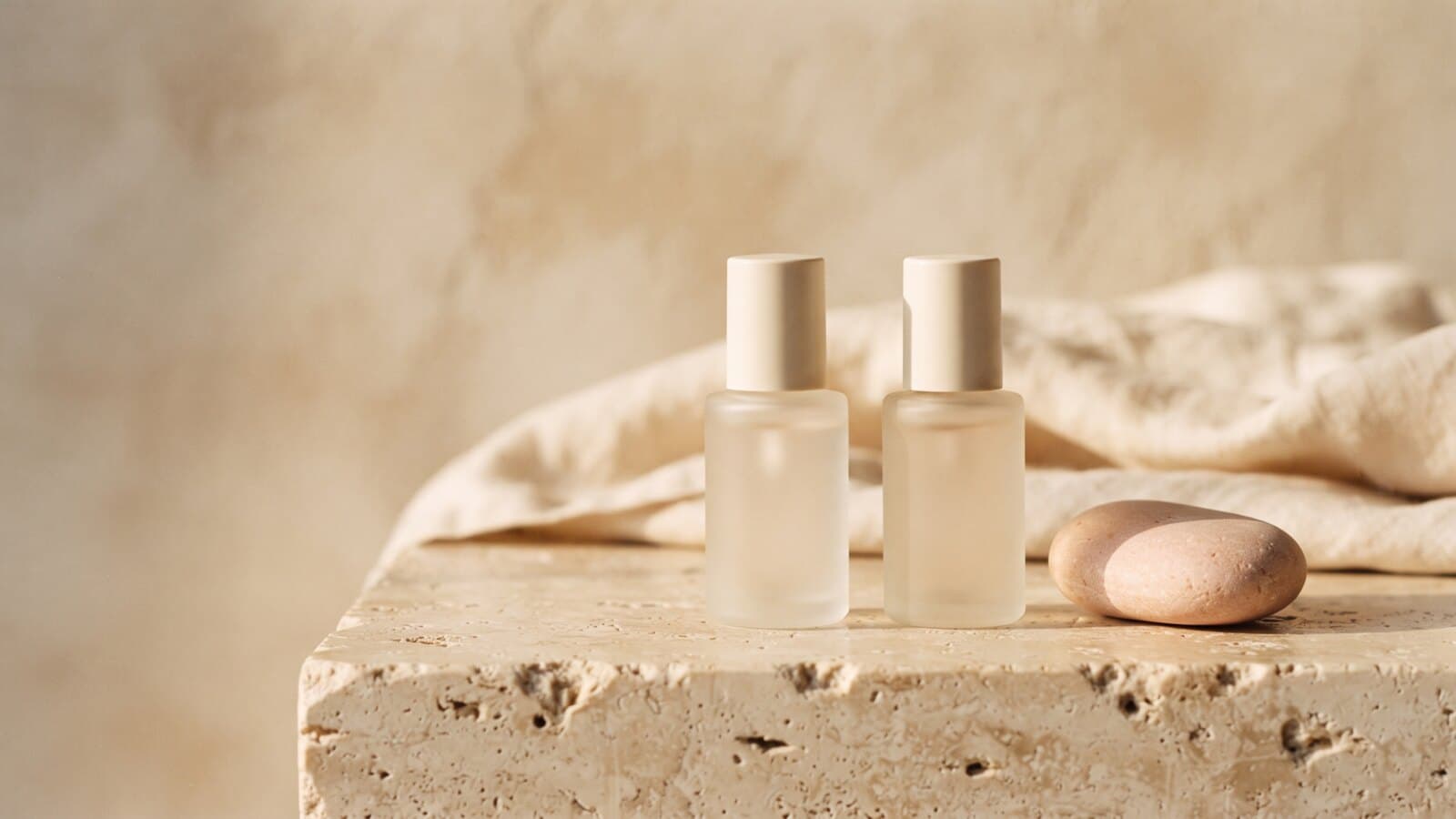 Two identical unlabeled frosted-glass serum bottles on a warm stone ledge in low side light, one beside a small pale pink river stone, a folded linen cloth behind.