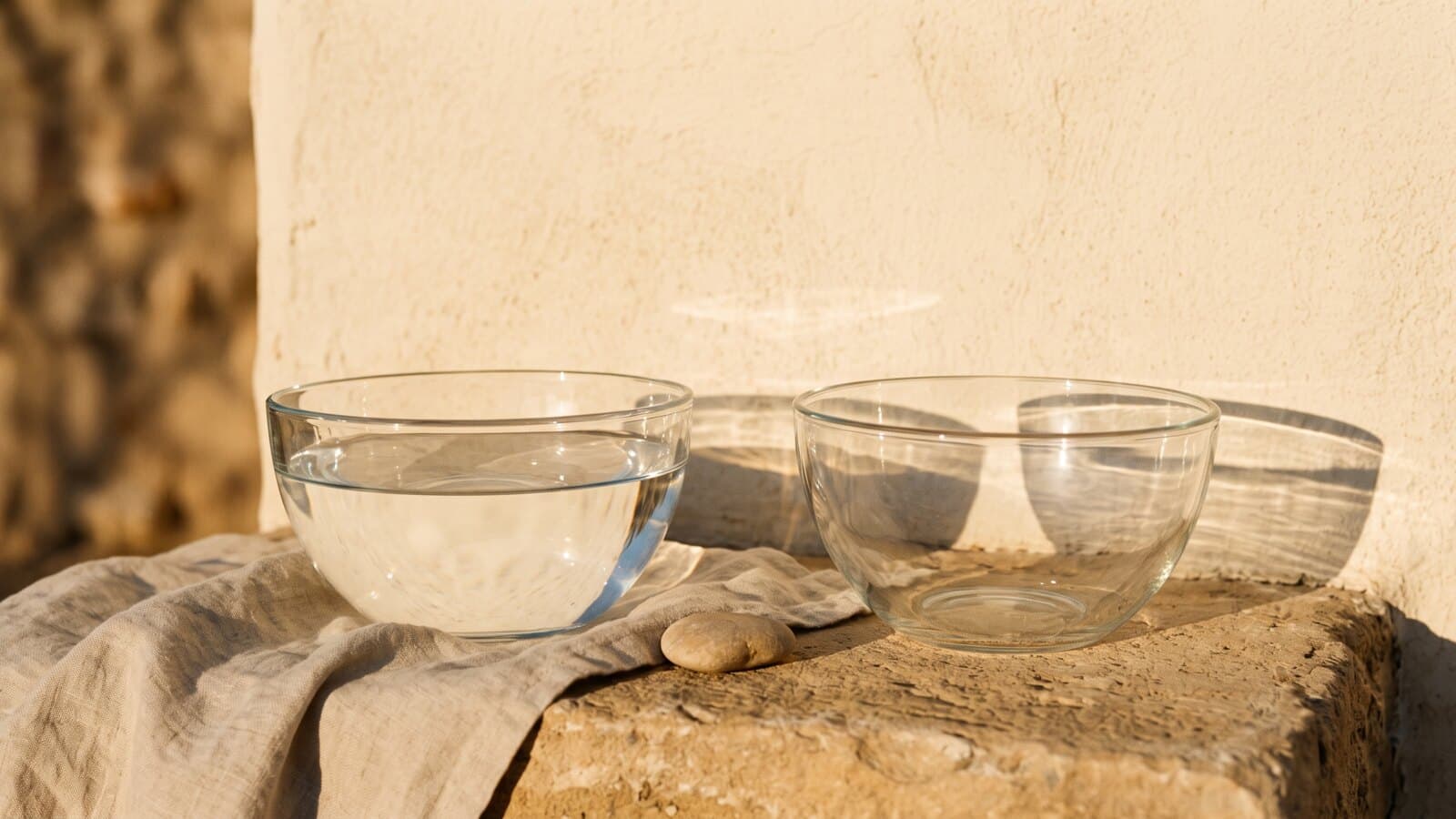Two identical clear glass bowls side by side on a stone ledge in low sun, one holding water, their different shadows falling on the plaster wall.
