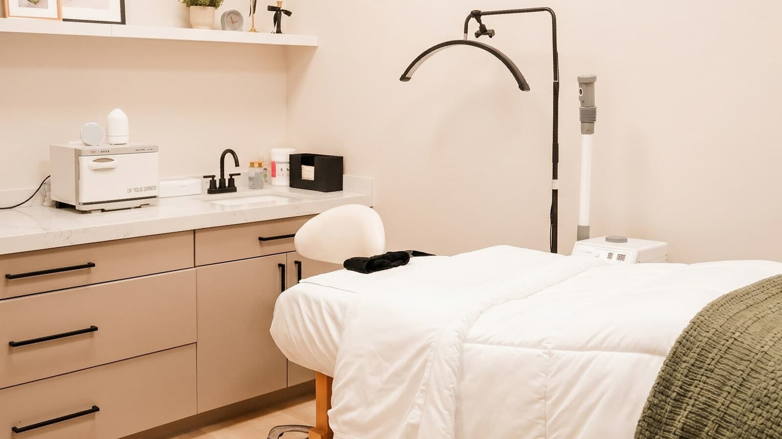 A treatment room at éclat: a made bed with a sage-green throw, soft lamplight on the shelf, and a curved treatment lamp waiting in the corner.