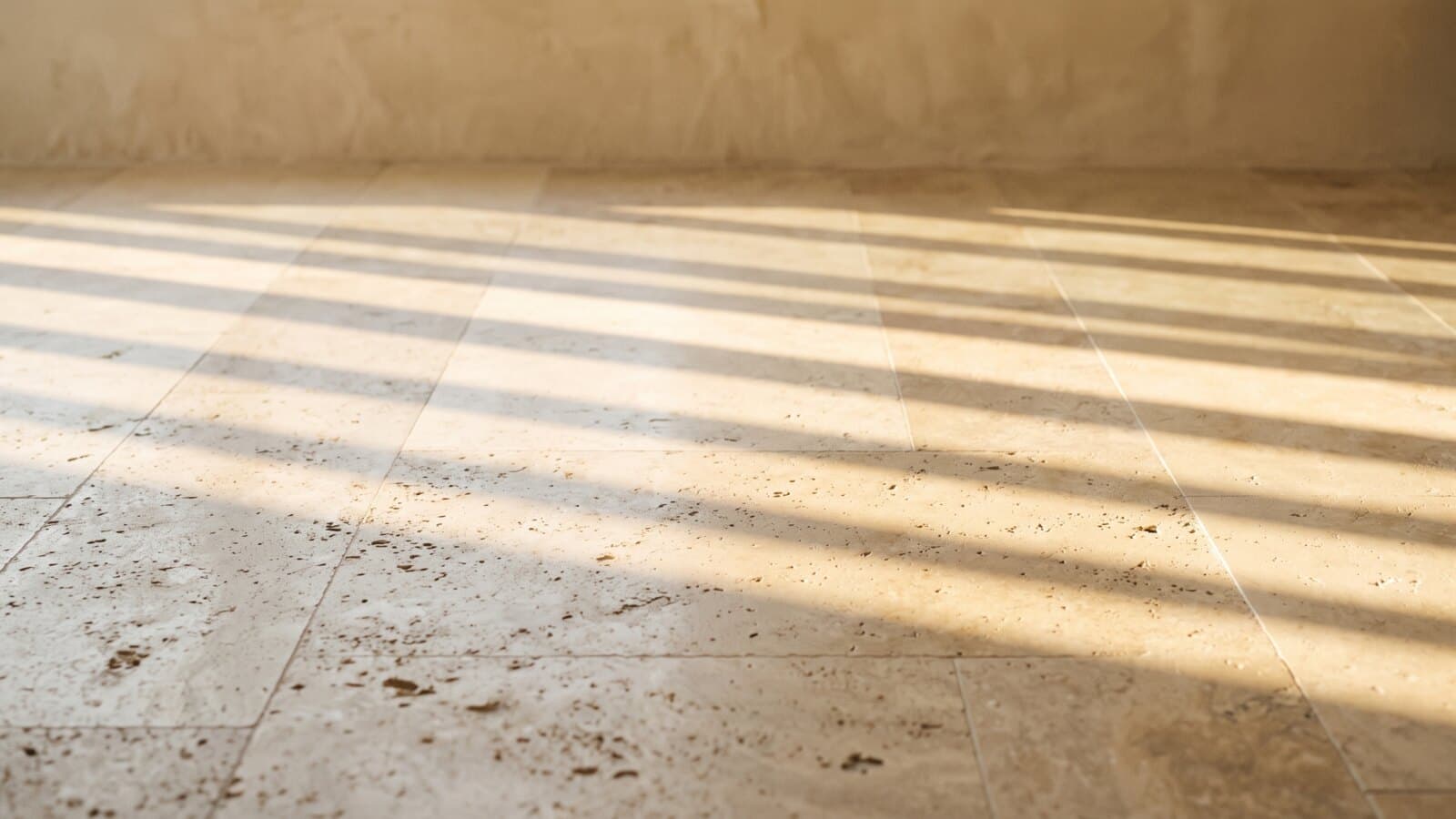 Bands of golden afternoon light and shadow from a tall window falling across a pale travertine floor.