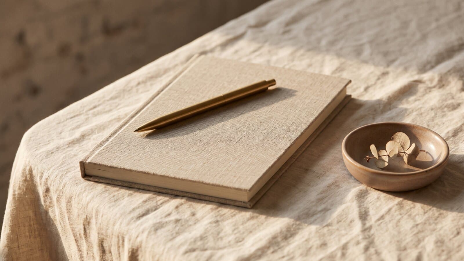 A closed linen-bound notebook with a brass pen resting on it beside a small ceramic dish holding a dried botanical, in low warm side light.
