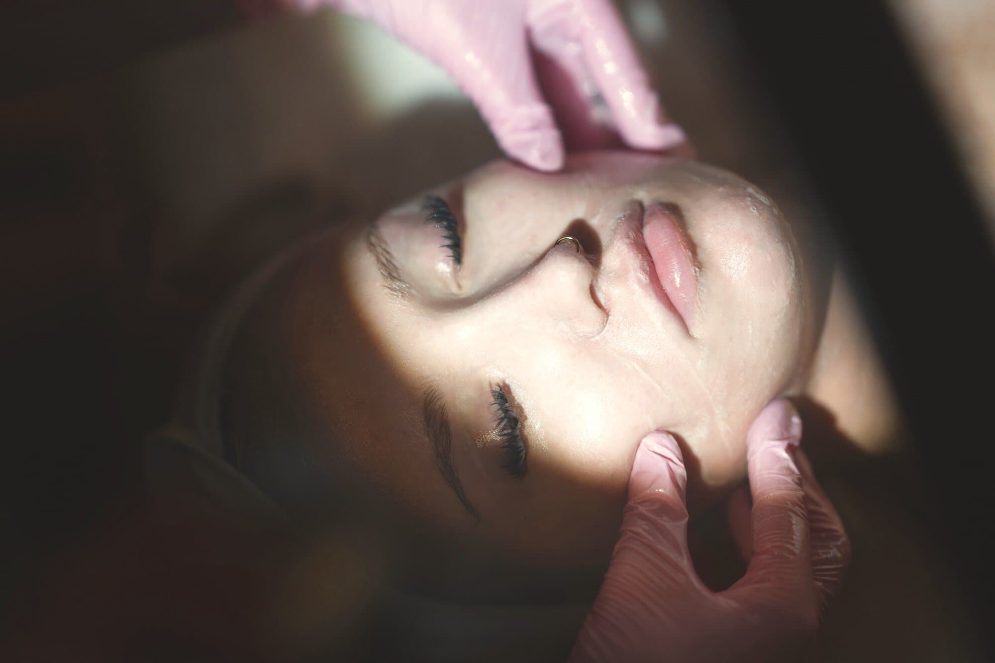 A provider's gloved hands working product across a guest's cheek and jaw