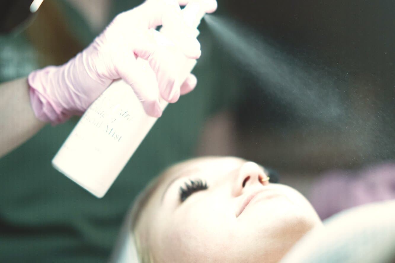 A gloved hand misting a guest's face under the treatment lamp